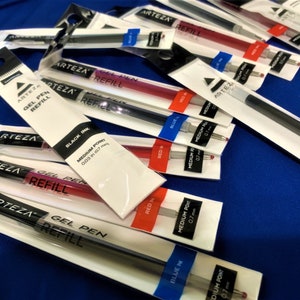 K&ouml;nnte beinhalten: Nahaufnahme von mehreren Packungen Arteza Gel-Stift-Nachf&uuml;llpatronen in verschiedenen Farben, darunter Schwarz, Rot und Blau. Die Packungen sind mit dem Markennamen, der Tintenfarbe und der Spitzengr&ouml;&szlig;e gekennzeichnet. Die Nachf&uuml;llpatronen haben eine mittlere Spitze von 0,7 mm.