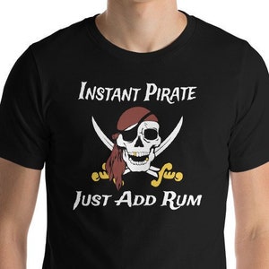 Op de afbeelding: Een zwart T-shirt met een witte schedel en gekruiste beenderen afbeelding. De schedel draagt een ooglapje en een rode bandana. De tekst "Instant Pirate Just Add Rum" is onder de afbeelding gedrukt.