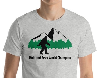 May include: A heather grey t-shirt with a black silhouette of Bigfoot walking in front of a green forest and snowy mountains. The text "Hide and Seek World Champion" is printed below the image.