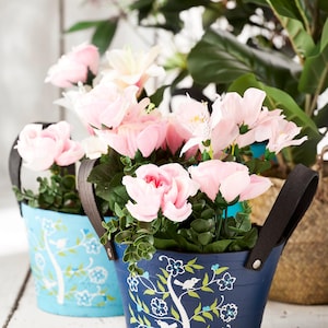 Puede incluir: Dos cubos decorativos de metal, uno azul claro y otro azul marino, llenos de flores rosas y vegetación. Los cubos tienen asas de cuero negro y un diseño de árbol y pájaro blanco. Los cubos están sobre una superficie de madera blanca.