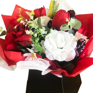 May include: A bouquet of red and white artificial flowers arranged in a black gift box. The flowers are wrapped in red tissue paper.
