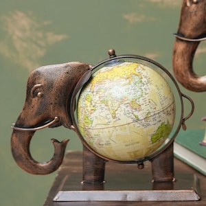 Peut inclure: Un ornement de table décoratif représentant un éléphant en métal de couleur bronze avec une finition texturée. Le corps de l'éléphant est intégré à un globe sphérique affichant une carte détaillée du monde dans des tons jaune et vert.