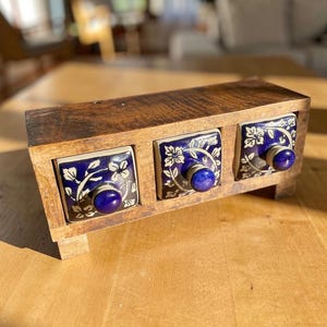 Puede incluir: Un especiero de madera con tres cajones. Cada cajón tiene un diseño floral azul y blanco con un pomo azul. El marco de madera tiene un acabado natural. El estante está elevado sobre pequeños pies de madera.
