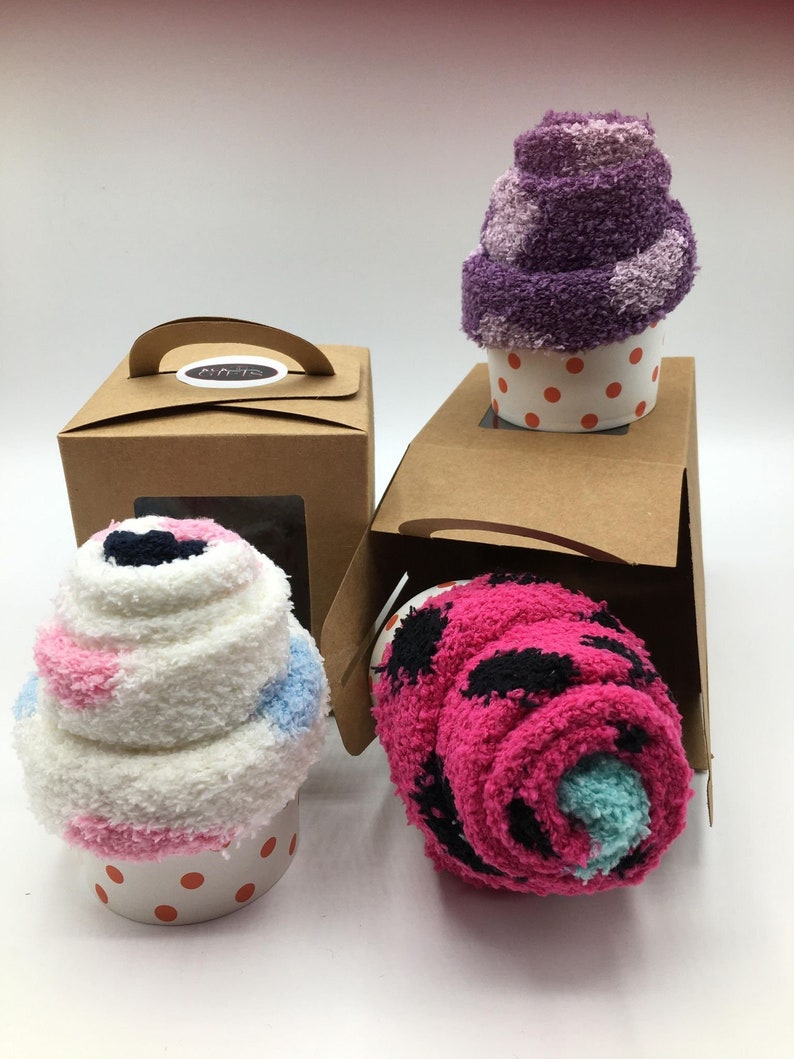 May include: Three cupcake-shaped socks in white, pink, blue, and purple, presented in white polka-dot cups. One is rolled up in a pink and black pattern. Two brown boxes with handles are in the background.