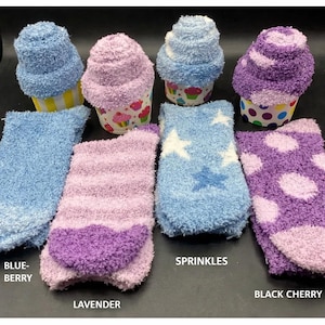 May include: Cozy, fluffy socks in various colors, including blue, lavender, and purple, are displayed. Some socks are rolled to resemble cupcakes. The socks feature patterns like stars and polka dots. Text on the image reads "Blueberry", "Lavender", "Sprinkles", and "Black Cherry".