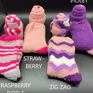 May include: A collection of fuzzy socks in various colors and patterns. The socks include shades of pink, purple, and gray, with designs such as stripes and zigzags. Text on the image reads "Raspberry Ripple", "Strawberry", "Zig Zag", and "Violet".