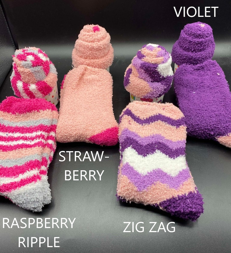 May include: A collection of fuzzy socks in various colors and patterns. The socks include shades of pink, purple, and gray, with designs such as stripes and zigzags. Text on the image reads "Raspberry Ripple", "Strawberry", "Zig Zag", and "Violet".