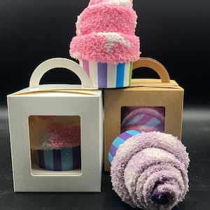 May include: A fluffy pink and white sock cupcake with a striped blue, purple, and green base. Two gift boxes with clear windows display similar sock cupcakes. One box is white, the other is brown. A single purple sock cupcake is also visible.