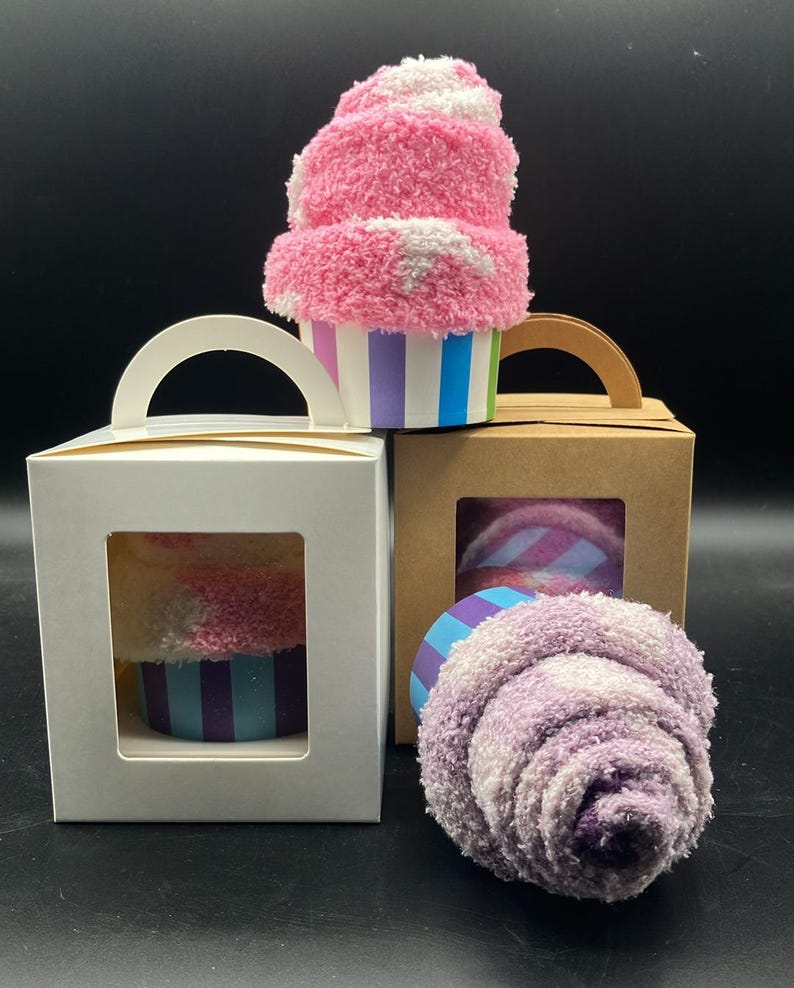 May include: A fluffy pink and white sock cupcake with a striped blue, purple, and green base. Two gift boxes with clear windows display similar sock cupcakes. One box is white, the other is brown. A single purple sock cupcake is also visible.