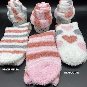 May include: Fluffy socks in three styles: Peach Melba, Black Forest, and Neapolitan. The socks are folded to resemble cupcakes and are displayed with unfolded pairs. The socks feature stripes and heart patterns in shades of pink, white, gray, and peach.