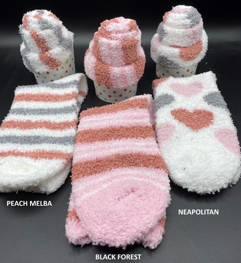 May include: Fluffy socks in three styles: Peach Melba, Black Forest, and Neapolitan. The socks are folded to resemble cupcakes and are displayed with unfolded pairs. The socks feature stripes and heart patterns in shades of pink, white, gray, and peach.