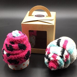 May include: Two fluffy sock cupcakes, one pink with black spots and the other white with pink, blue, and black stripes, sit on a dark surface. A brown gift box with a handle and a window displays a cupcake. The cupcakes are in paper cups.