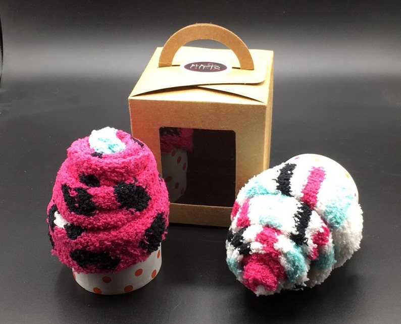 May include: Two fluffy sock cupcakes, one pink with black spots and the other white with pink, blue, and black stripes, sit on a dark surface. A brown gift box with a handle and a window displays a cupcake. The cupcakes are in paper cups.