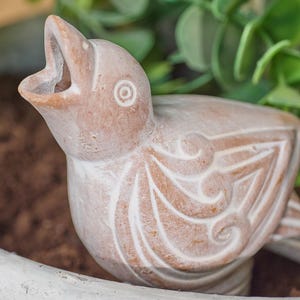 Puede incluir: Primer plano de una pequeña escultura de pájaro de color terracota con el pico abierto. El pájaro tiene detalles blancos y tallados en su cuerpo. La escultura se encuentra sobre un fondo borroso de follaje verde y una superficie blanca.
