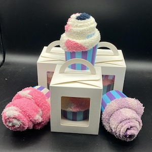 May include: A cupcake-shaped gift set featuring fluffy, colorful socks. The socks are designed to resemble a cupcake with white, pink, and blue accents. They are presented in white gift boxes with clear windows and handles, perfect for gifting.