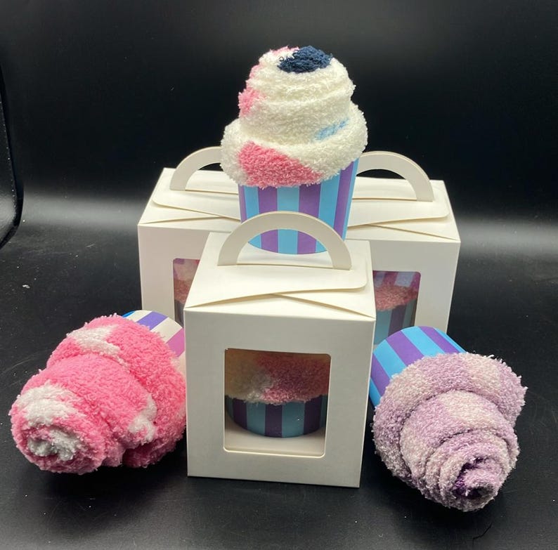 May include: A cupcake-shaped gift set featuring fluffy, colorful socks. The socks are designed to resemble a cupcake with white, pink, and blue accents. They are presented in white gift boxes with clear windows and handles, perfect for gifting.