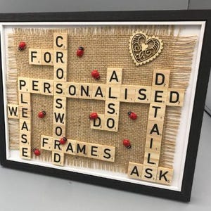 Może przedstawiać: Oprawiona dekoracja ścienna z drewnianymi kafelkami z literami ułożonymi w krzyżówkę na tle z juty. Tekst brzmi: FOR CROSSWORD PERSONALISED DETAILS PLEASE DO FRAMES ASK. Zawiera małe czerwone biedronki oraz drewniane serce jako elementy ozdobne.