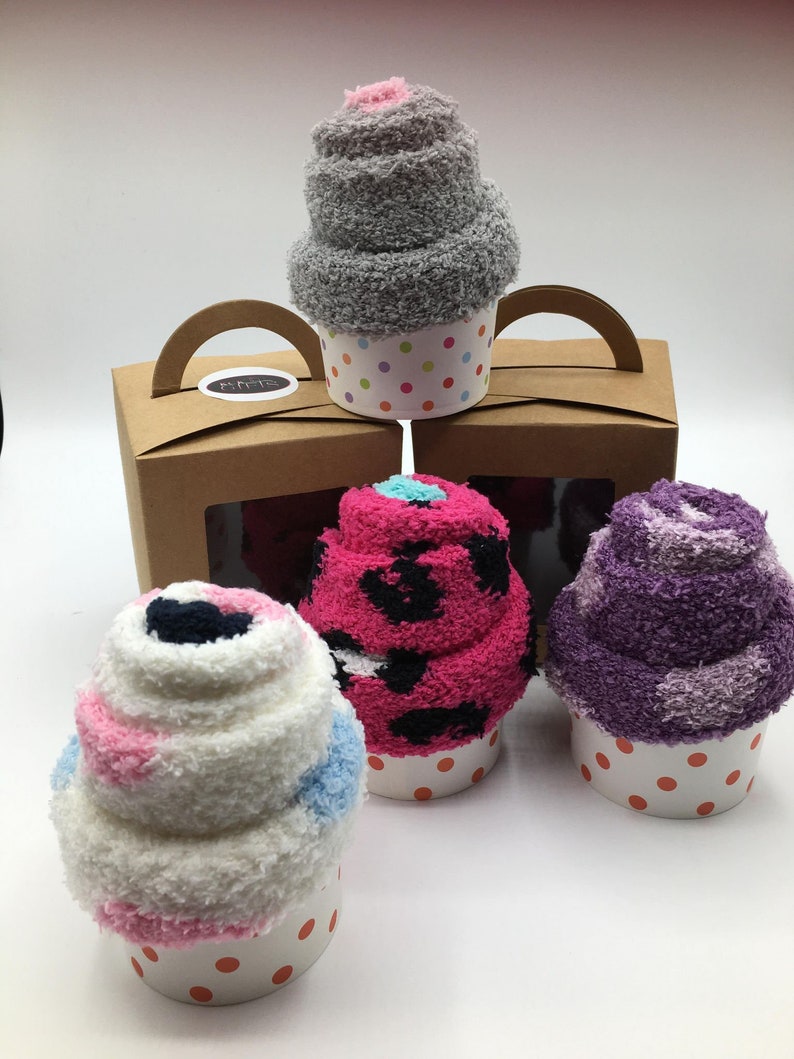 May include: A collection of colorful sock cupcakes, including gray, pink, purple, and white, each in a polka-dot cupcake liner. A brown box with a handle and a window displays more sock cupcakes.
