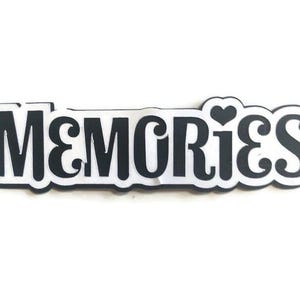 May include: A black and white sticker with the word "MEMORIES" in a bold, rounded font. The word is outlined in white, with a small heart replacing the dot over the "i". The sticker is on a white background.