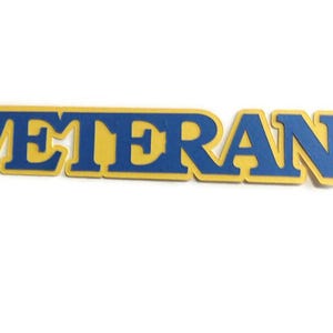 May include: A blue and yellow wooden sign that spells out the word "VETERAN". The letters are blue with a yellow outline. The sign is on a white background.
