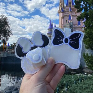 May include: Two white fabric keychains with black and white embroidery. One keychain features a bow and the other features a bow and a Mickey Mouse head. The keychains are held in front of a pink and blue castle.