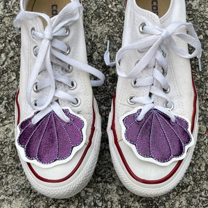 May include: A pair of white canvas trainers with purple glitter seashell patches on the toes. The laces are white and tied in a bow. The trainers have red trim around the edges.