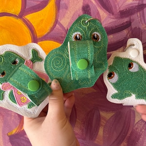 May include: Three green felt frog keyrings with embroidered details. Each keyring has a different design, including a frog with a crown, a frog with a flower, and a frog with a simple design. The keyrings are attached to white felt backing.