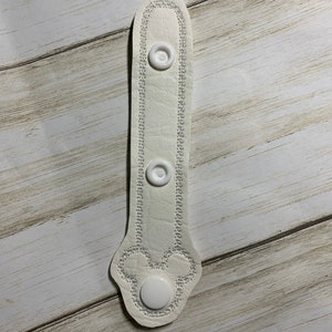 May include: White fabric strap with three white plastic buttons. The strap has a decorative stitch around the edges and a Mickey Mouse design at the bottom.