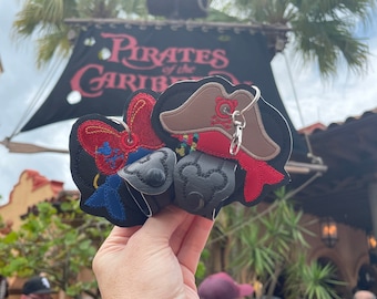 May include: A pair of black felt Mickey Mouse ears with a red and blue pirate hat and a brown pirate hat with a skull and crossbones design. The ears are attached to a silver keyring.