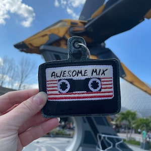 May include: A black and white cassette tape keychain with red and white stripes. The keychain says "Awesome Mix" in white lettering. The keychain is being held in front of a large, yellow and gray spaceship.