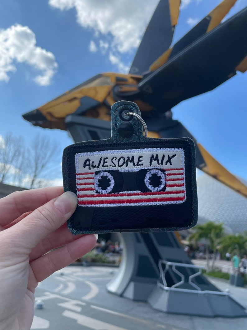 May include: A black and white cassette tape keychain with red and white stripes. The keychain says "Awesome Mix" in white lettering. The keychain is being held in front of a large, yellow and gray spaceship.