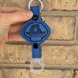 May include: Blue leather keychain with a black silhouette of a couple in a boat on a cloud. The keychain has a clear plastic bottle opener attached.