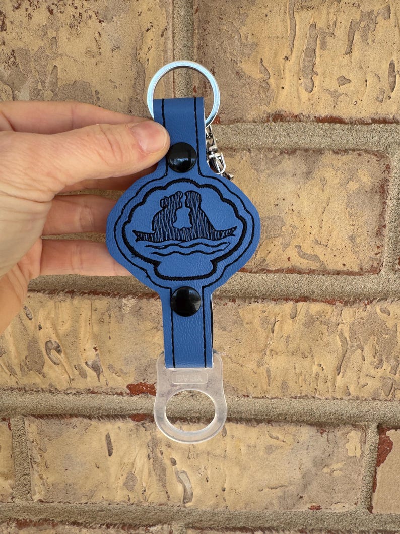 May include: Blue leather keychain with a black silhouette of a couple in a boat on a cloud. The keychain has a clear plastic bottle opener attached.