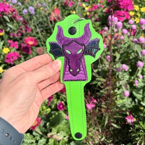 May include: A neon green key fob featuring a purple dragon design with black accents. The dragon has horns and wings, and the fob has a metal ring for attaching keys. The background is a colorful garden.