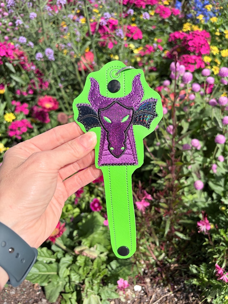May include: A neon green key fob featuring a purple dragon design with black accents. The dragon has horns and wings, and the fob has a metal ring for attaching keys. The background is a colorful garden.