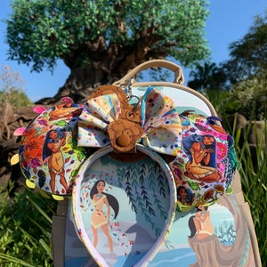 May include: A themed ear headband with a matching backpack. The headband features colorful fabric ears with a Pocahontas design and a large bow. The backpack has a matching design with a Pocahontas illustration. The background includes a large tree and blue sky.