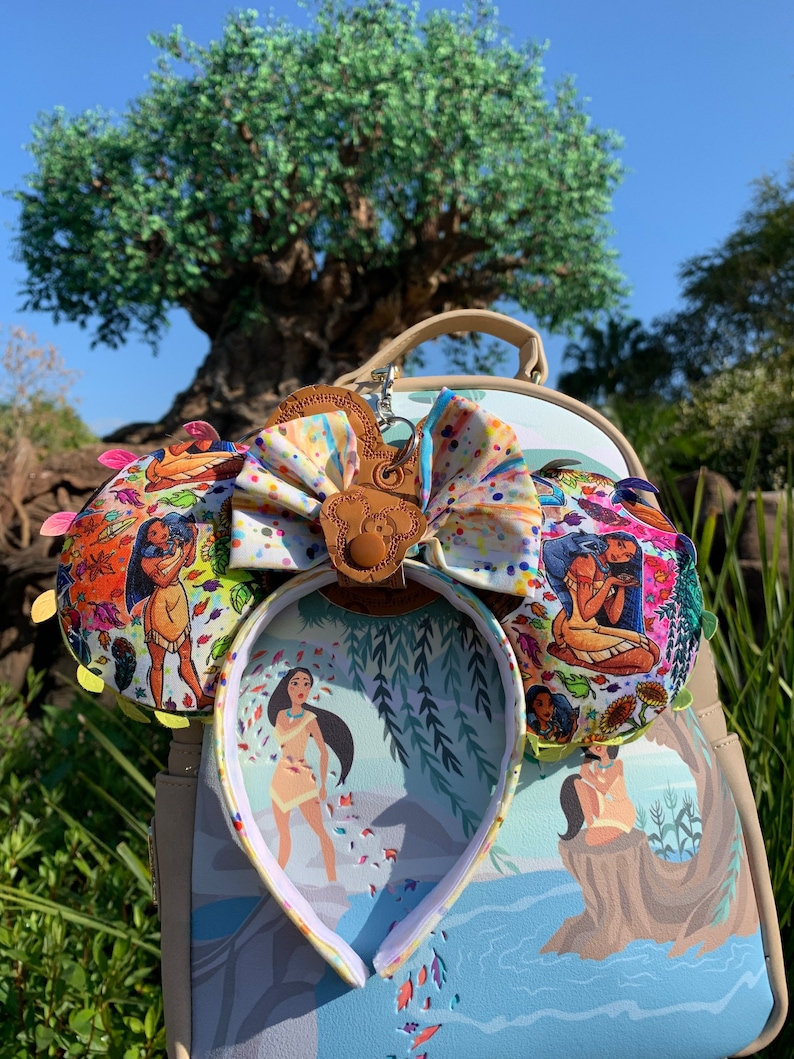 May include: A themed ear headband with a matching backpack. The headband features colorful fabric ears with a Pocahontas design and a large bow. The backpack has a matching design with a Pocahontas illustration. The background includes a large tree and blue sky.