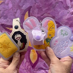 May include: Four embroidered keychains featuring Disney characters. The keychains are purple, yellow, and white with embroidered details. The characters include Rapunzel, a frog, a sun, and a cloud.
