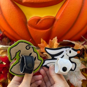 May include: Two embroidered keychains featuring characters from the Nightmare Before Christmas. One keychain is green and features a ghost with a black leather strap. The other keychain is white and features Zero, the ghost dog, with a black button.