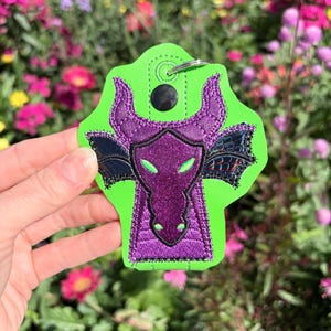 May include: A green and purple dragon keychain. The keychain features a dragon design with a purple face, horns, and wings, set against a bright green background. The dragon's eyes are green, and the wings are black.
