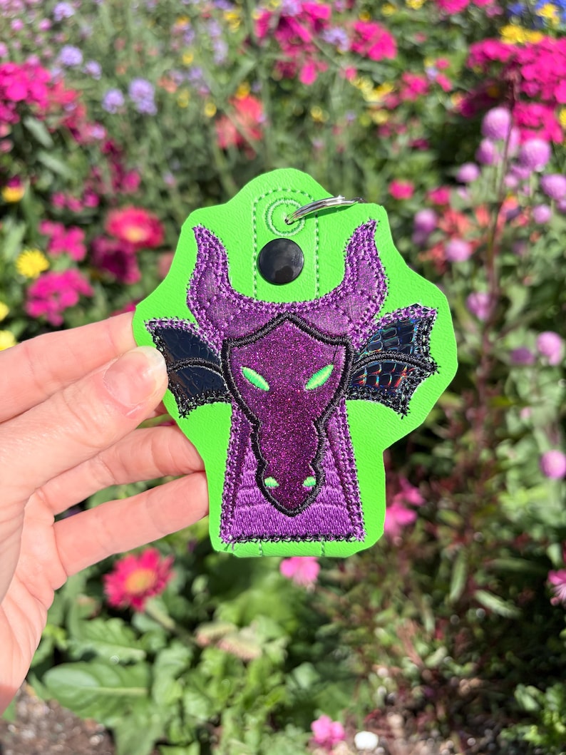 May include: A green and purple dragon keychain. The keychain features a dragon design with a purple face, horns, and wings, set against a bright green background. The dragon's eyes are green, and the wings are black.