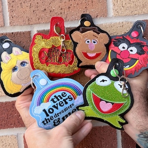 May include: A collection of embroidered keychains featuring characters from The Muppet Show. The keychains include Miss Piggy, Kermit the Frog, Fozzie Bear, Animal, and a keychain with the text "the lovers the dreamers & me". The keychains are in various colors.