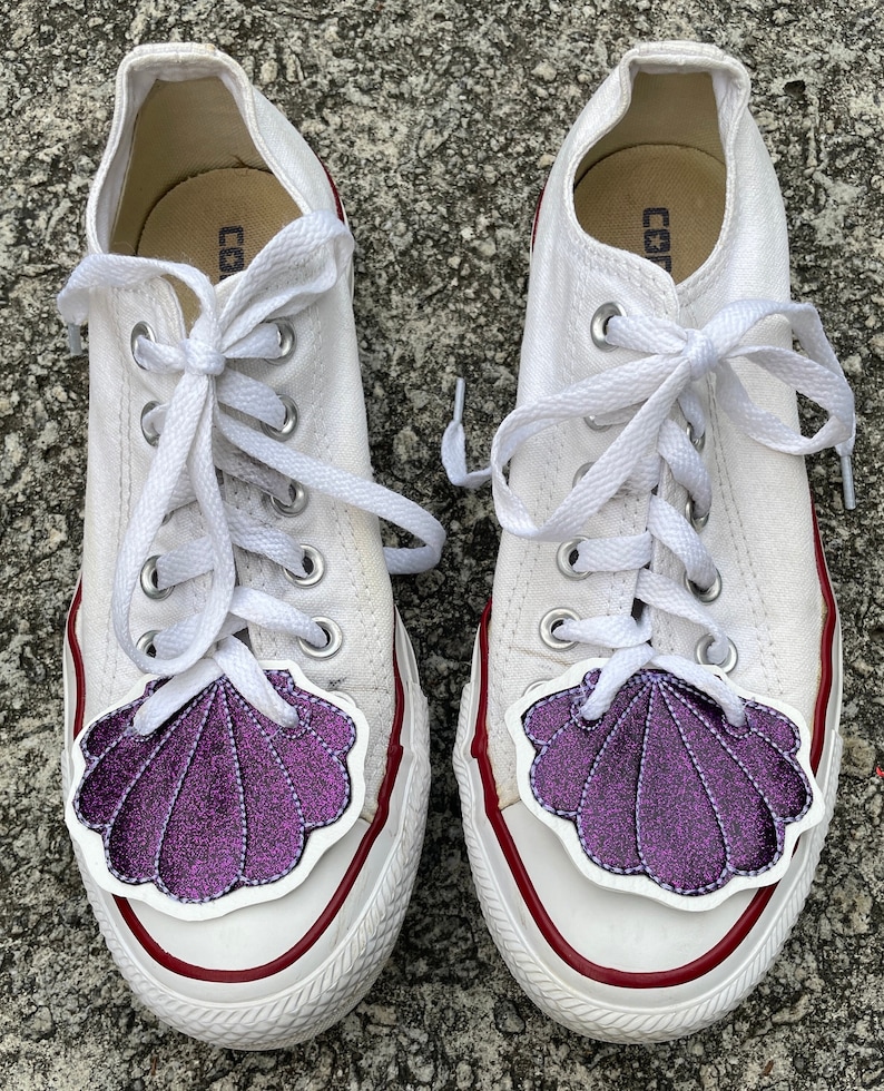 May include: A pair of white canvas sneakers with purple glitter seashell patches on the toes. The laces are white and tied in a bow. The sneakers have red trim around the edges.