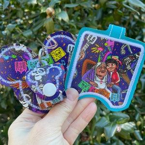 Puede incluir: Dos llaveros de tela morado y blanco con un estampado colorido de personajes de la película Coco. El llavero de la izquierda tiene forma de orejas de Mickey Mouse. El llavero de la derecha es de forma rectangular con esquinas redondeadas. Ambos llaveros tienen un borde blanco.