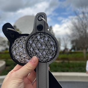 May include: Two gray and black fabric keychains with a white embroidered design of a sphere with a grid pattern. The keychains are attached to silver key rings.