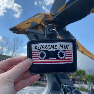 May include: A black and white cassette tape keychain with the text "Awesome Mix" embroidered on it. The keychain is being held in front of a large, futuristic-looking spaceship.
