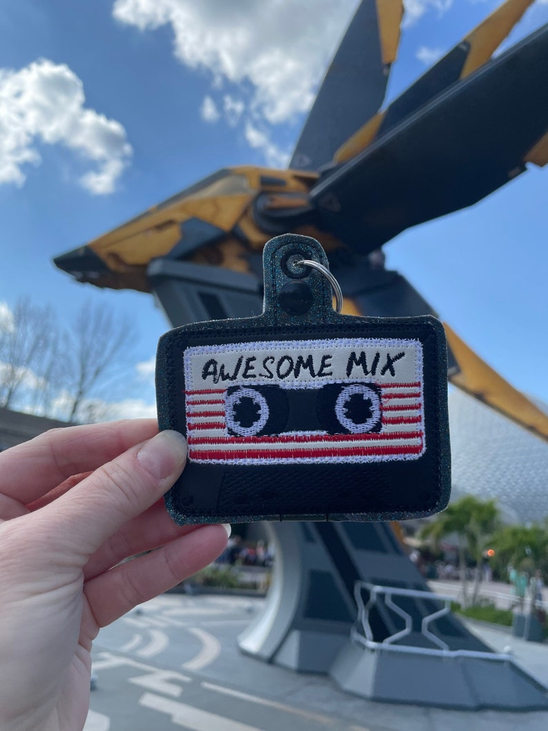 May include: A black and white cassette tape keychain with the text "Awesome Mix" embroidered on it. The keychain is being held in front of a large, futuristic-looking spaceship.