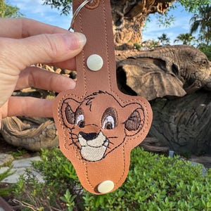 May include: Brown leather keychain featuring an embroidered image of Simba from The Lion King. The keychain has a white button and a metal ring.