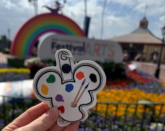 May include: A white Mickey Mouse-shaped keyring with a paint palette and paintbrushes. The keyring is in front of a rainbow arch and the words "Festival of the Arts".