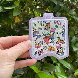 May include: A purple and white fabric patch with a repeating pattern of a cartoon duck holding a potted plant. The duck is wearing a blue dress and a pink bow. The patch has a white border and a purple loop at the top.